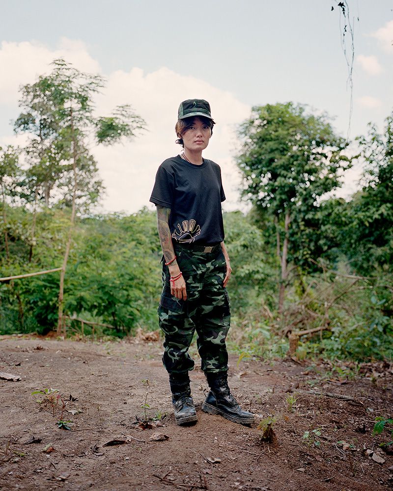 A young Myanmar soldier is standing outside wearing an army hat, pants, boots, and a t-shirt.
