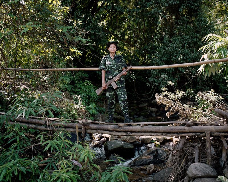 A young Myanmar soldier stands on a bridge with a machine gun in their arms.