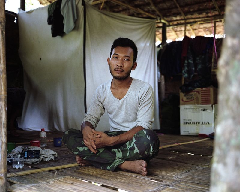 A Myanmar soldier sits on the floor inside a hut with his arms and legs crossed and a communications device next to him.
