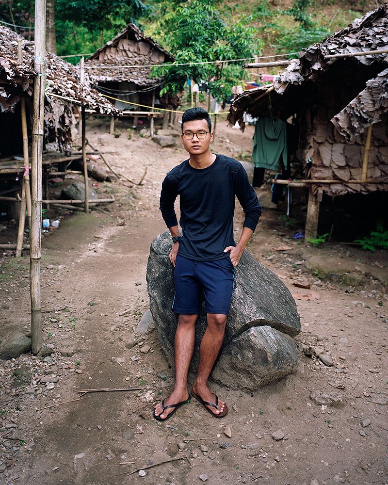 A young Myanmar man leans against a boulder with his left hand in the pocket with three huts in the background.