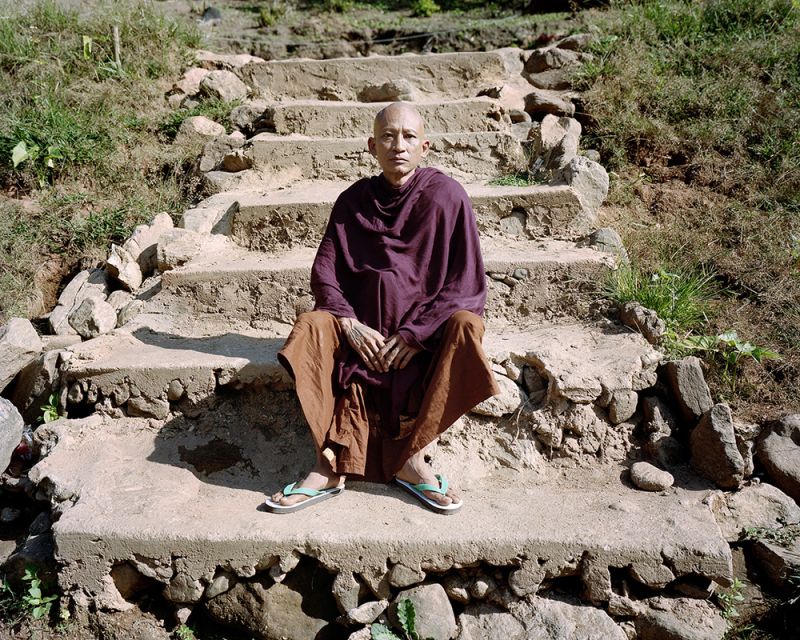 A man photographed sitting on the stairs outside, wearing different-coloured robes and flip-flops.