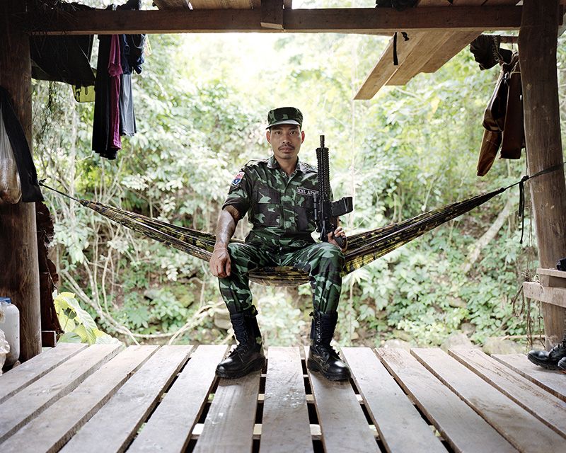 A Myanmar soldier is posing for a portrait sitting in a military hummock on a deck. 