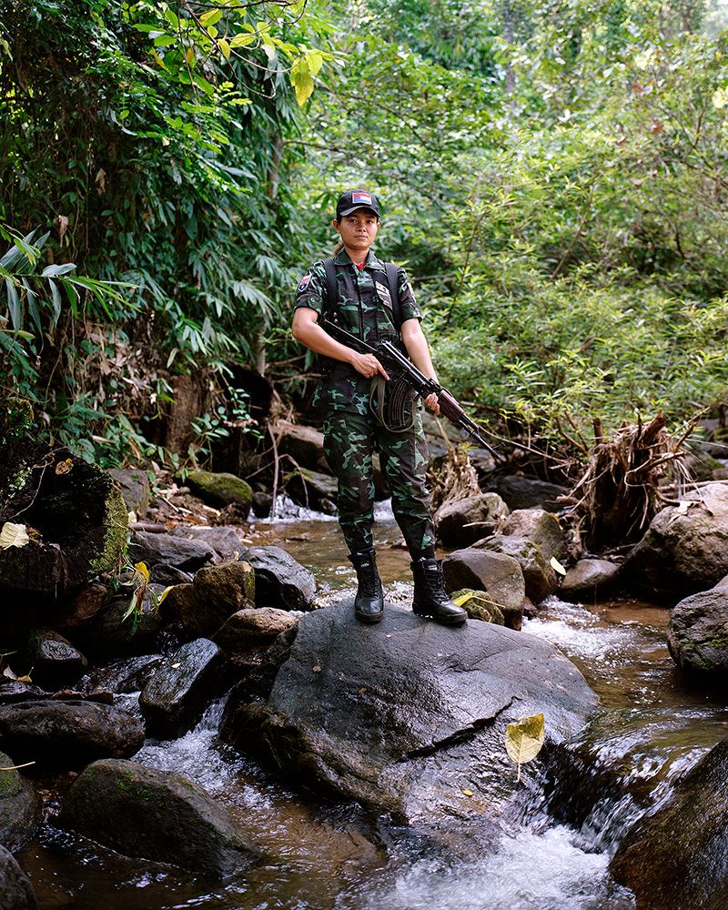 A young Myanmar soldier is posing for a portrait, standing on a boulder in the middle of a creek holding a machine gun. 