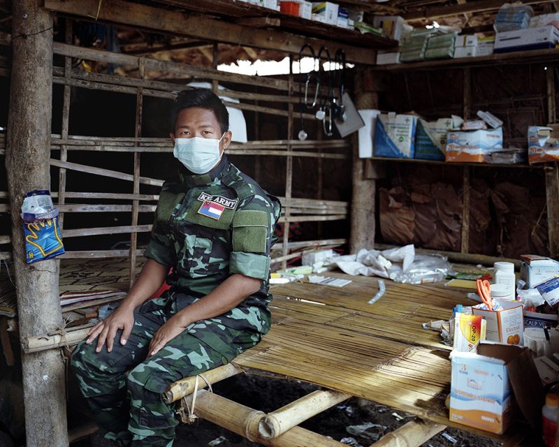 A young Myanmar soldier is posing for a portrait sitting on a first aid table, wearing a medical mask, surrounded by medical supplies.