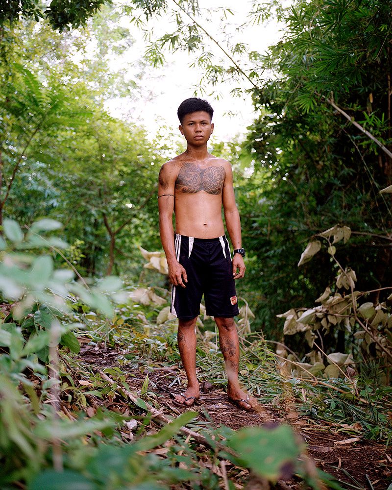 A young Myanmar man with a tattooed body posing in the middle of a forest wearing basketball shorts and flip-flops.