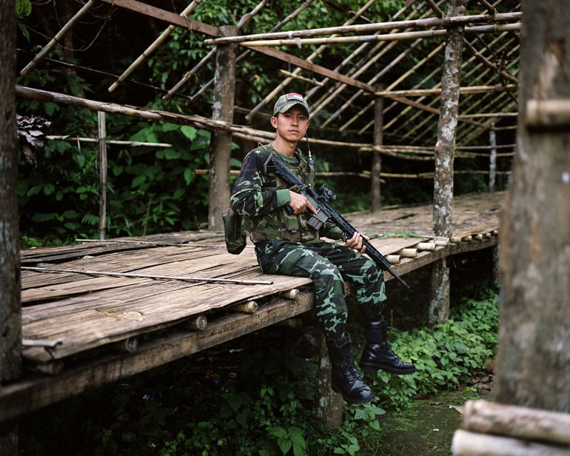 A young Myanmar soldier is sitting on a deck with a machine gun in his arms.