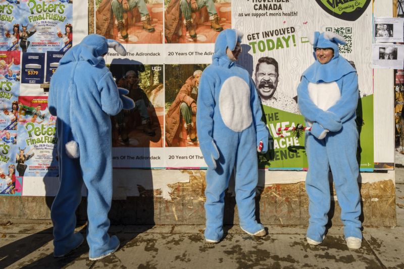 Three people dressed as blue bunnies in front of a wall covered in various posters on a sunny day.