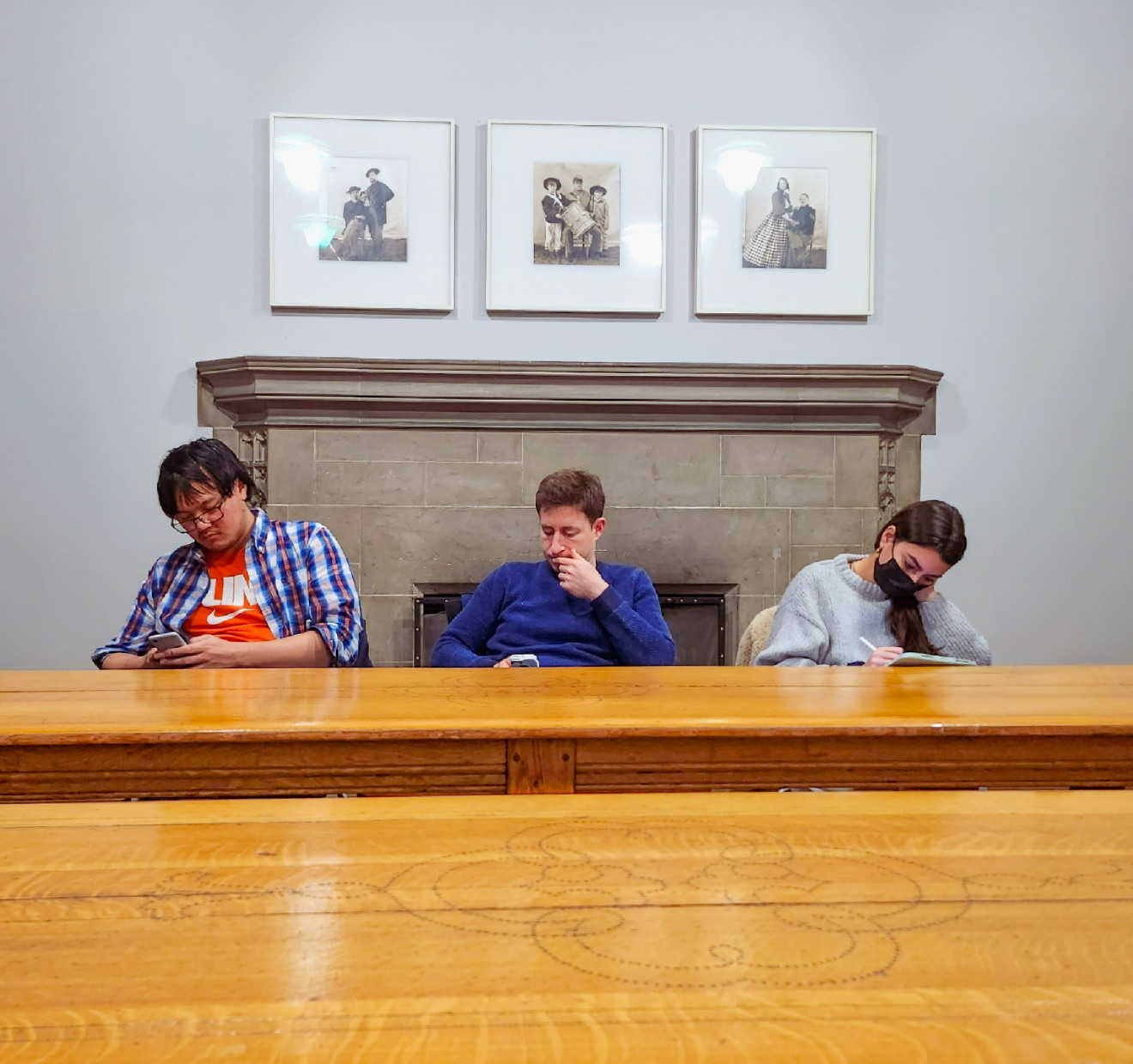 Three students sitting behind a desk and in front of a fireplace with three framed photographs above it, engaging with their cellular devices.
