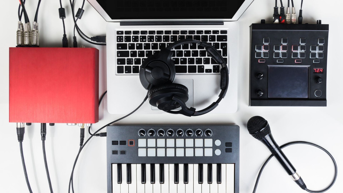 A picture of an open mac laptop with headphones on top of it, a red interface to the left of it, a beat machine to the right of it, and a mini keyboard just below the laptop