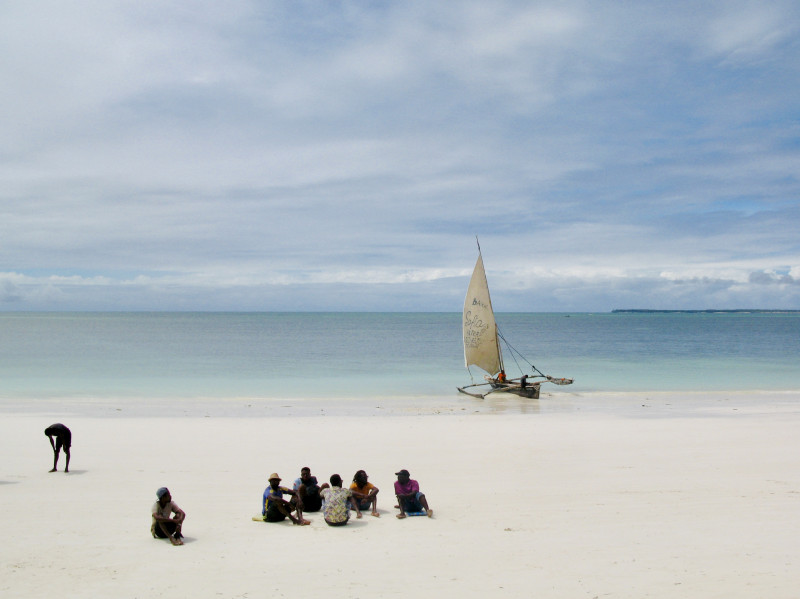 A group of people sitting on a sand beach with a sailing boat in the background. 