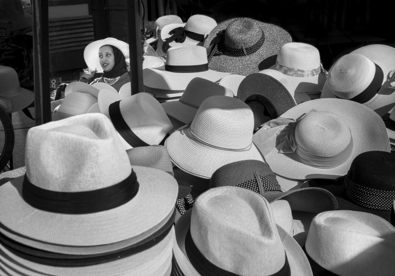 A display of straw hats and a woman's reflection in the window as she is trying one of the hats. 