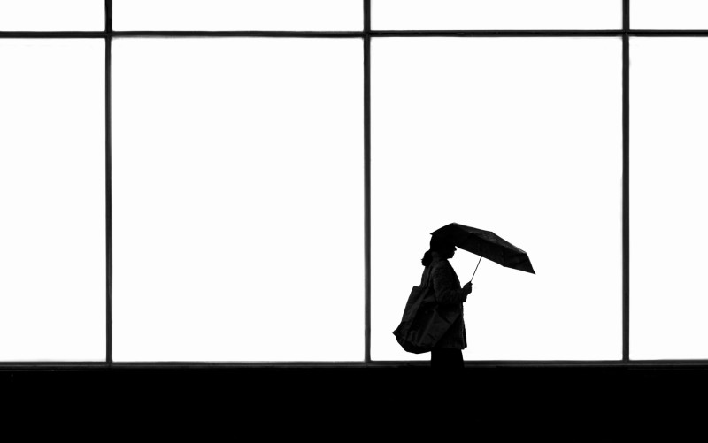A silhouette of a woman walking by a window with an open umbrella and a bag in her hand. 
