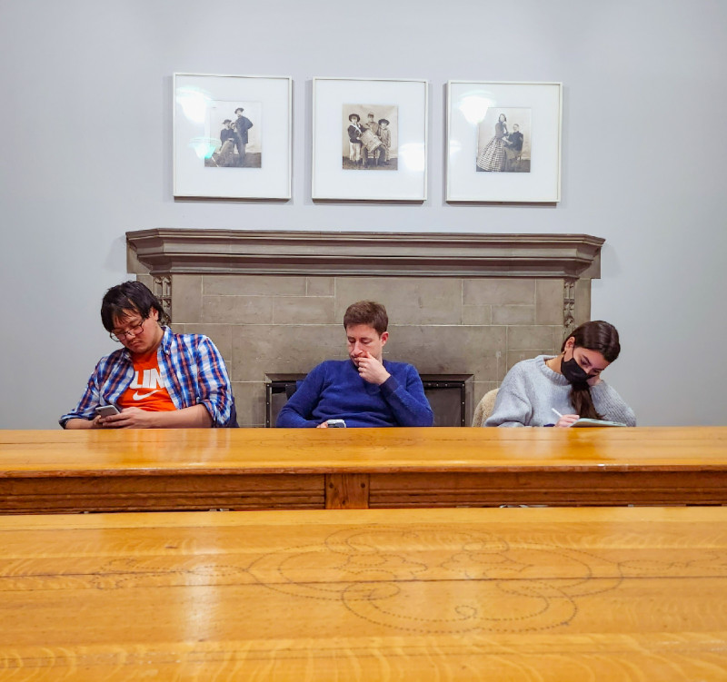 Three students seated behind a desk and in front of a fireplace. ABove the fireplace are three black and white framed images. The students are engaged in looking at their cellphones and writing. 