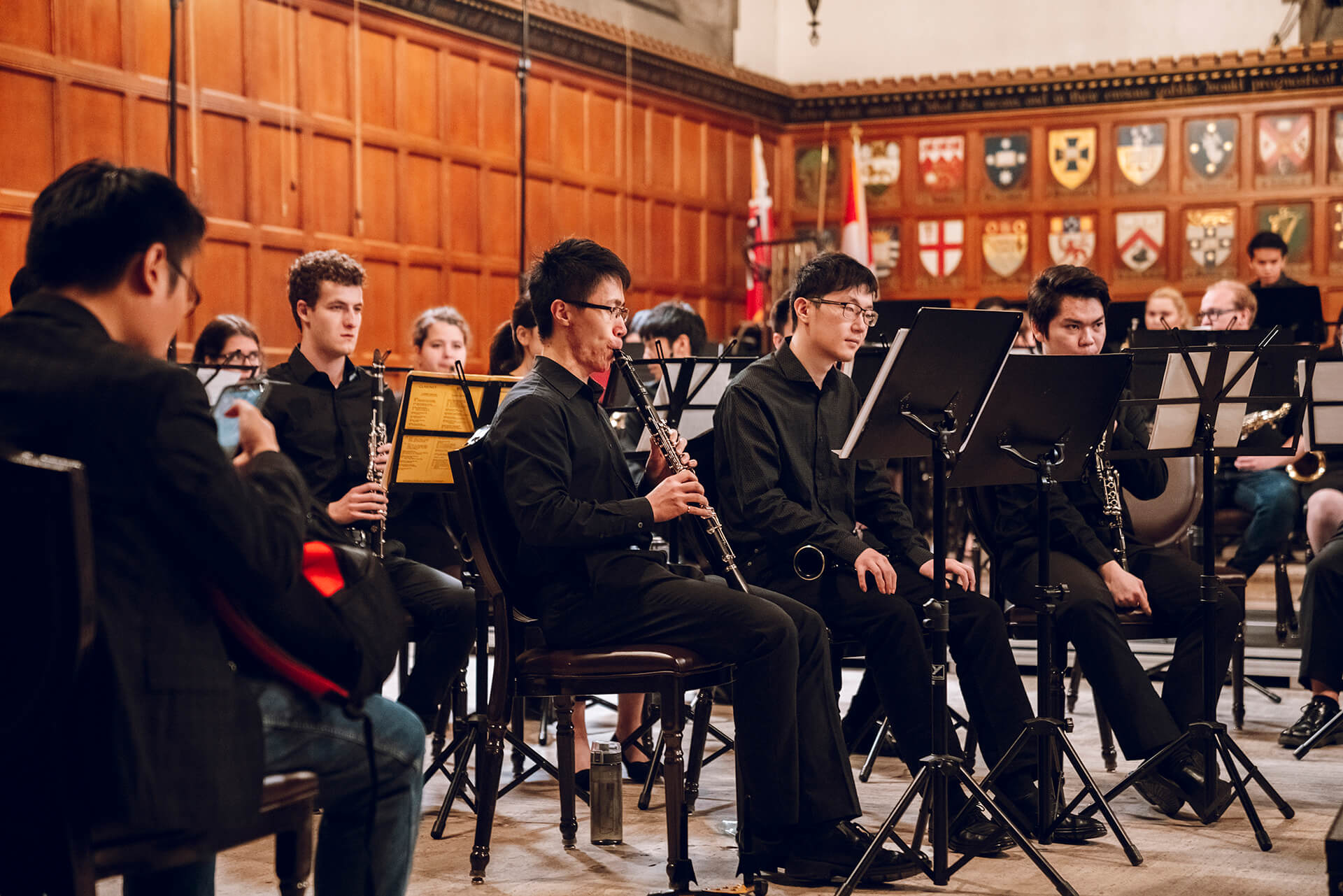 Band sitting with their musical instruments and a large collection of heraldry shields display on the wall beside them