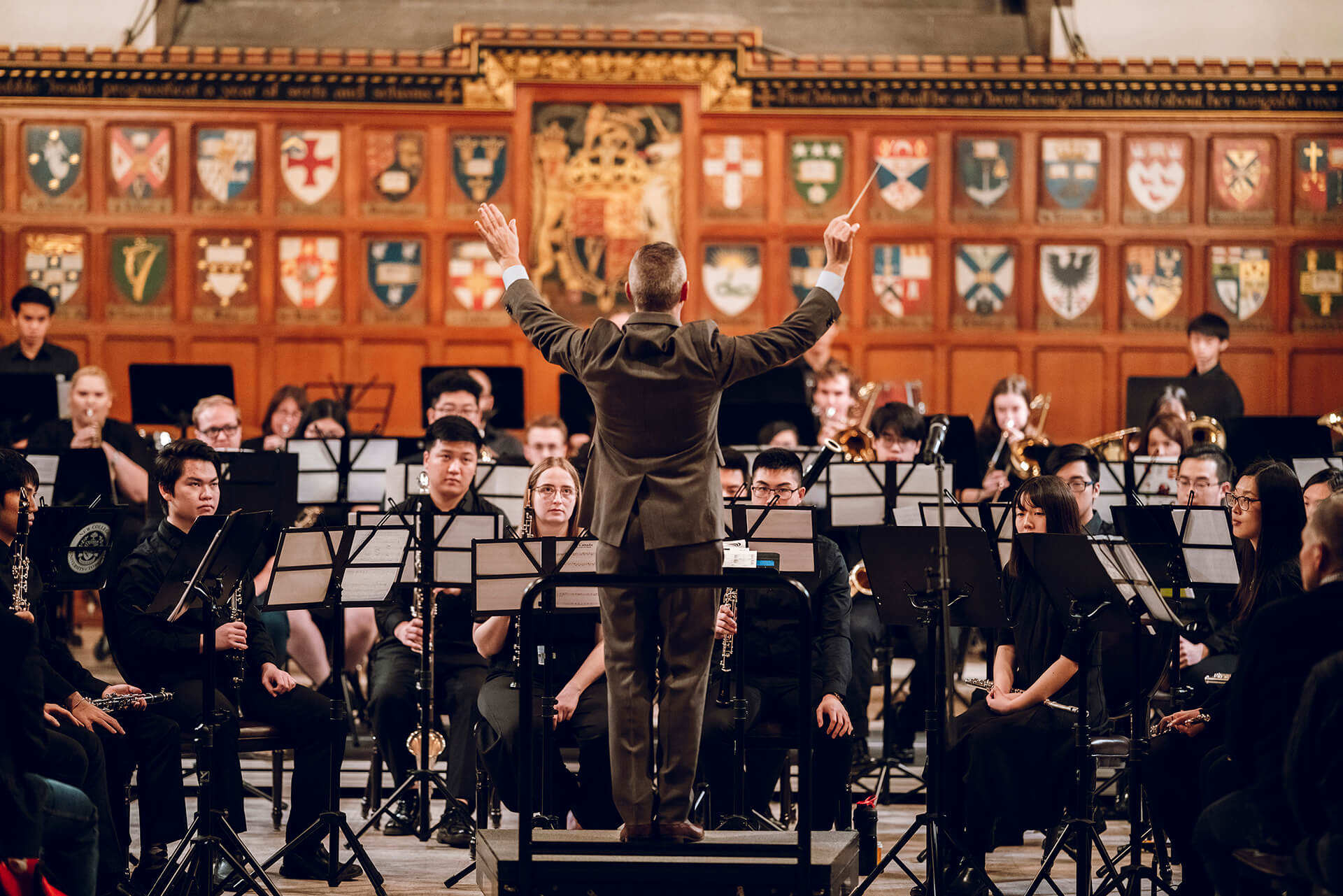 Band sitting with their musical instruments and a large collection of heraldry shields display on the wall behind them and a conductor standing and leading the band