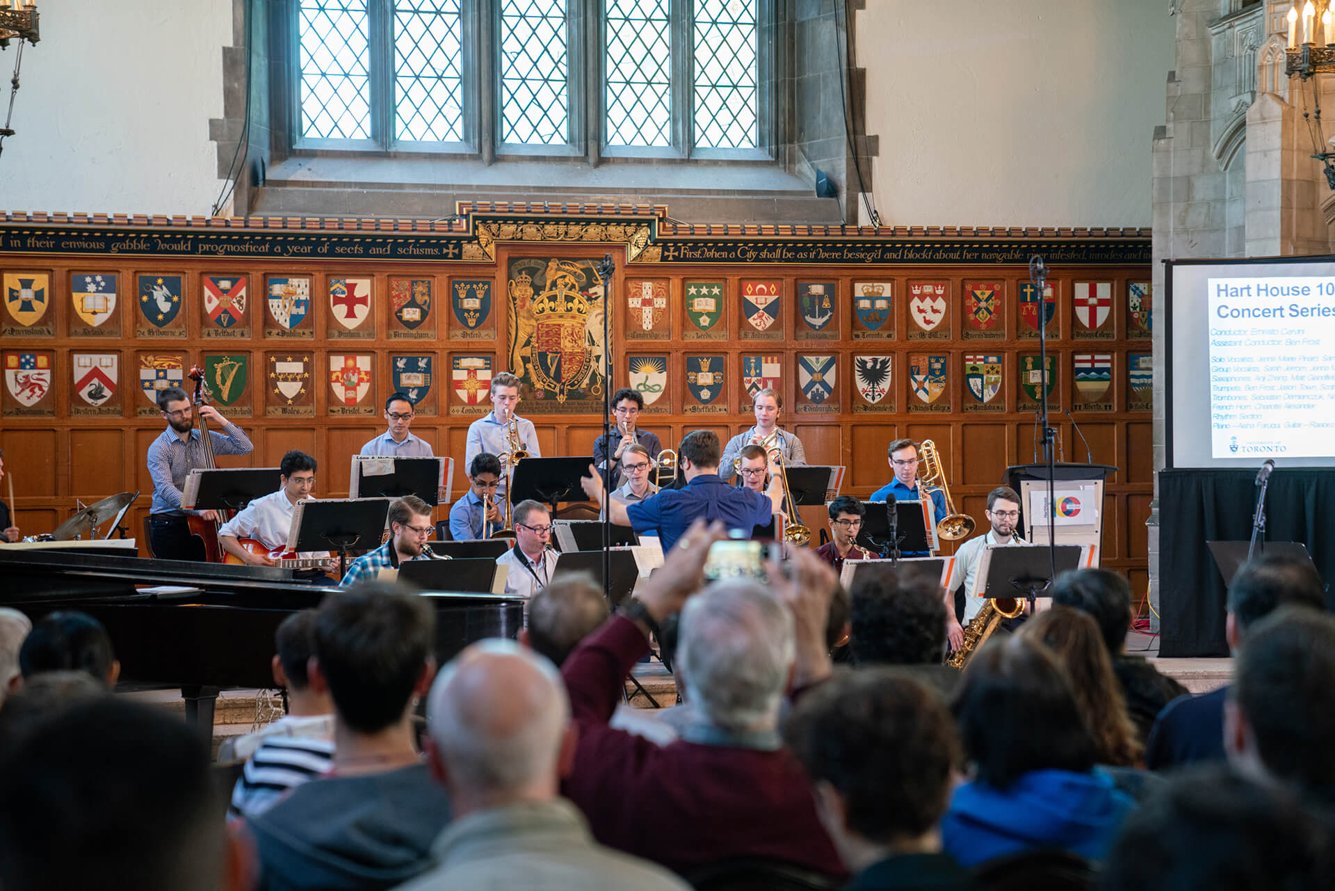 Band sitting and playing their musical instruments for sitting audience and a conductor standing conducting the band a large collection of of heraldry shields displays on the wall behind them