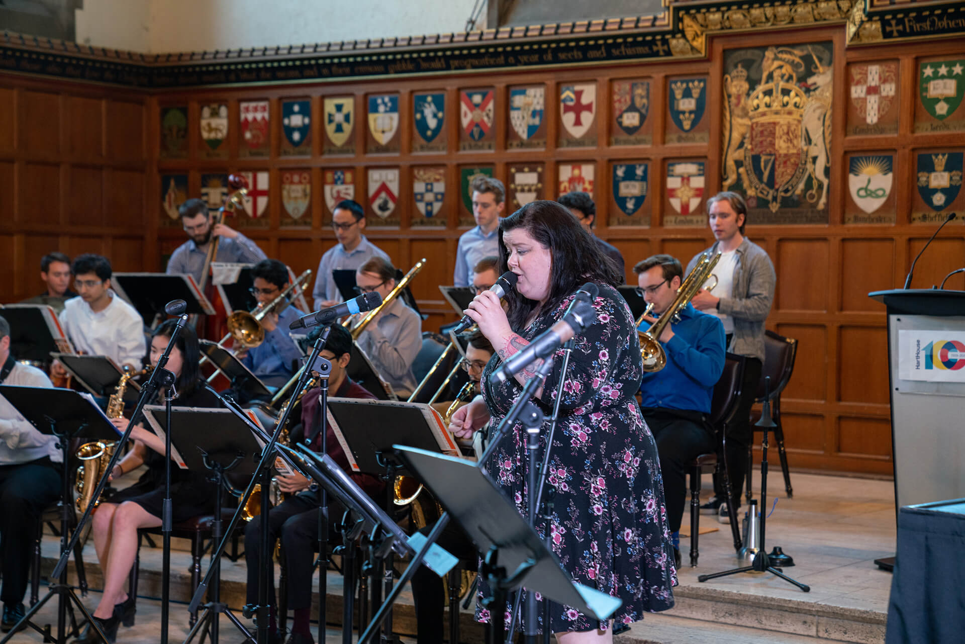 Band sitting and playing their musical instruments a woman standing and singing a large collection of heraldry shields displays on the wall behind them