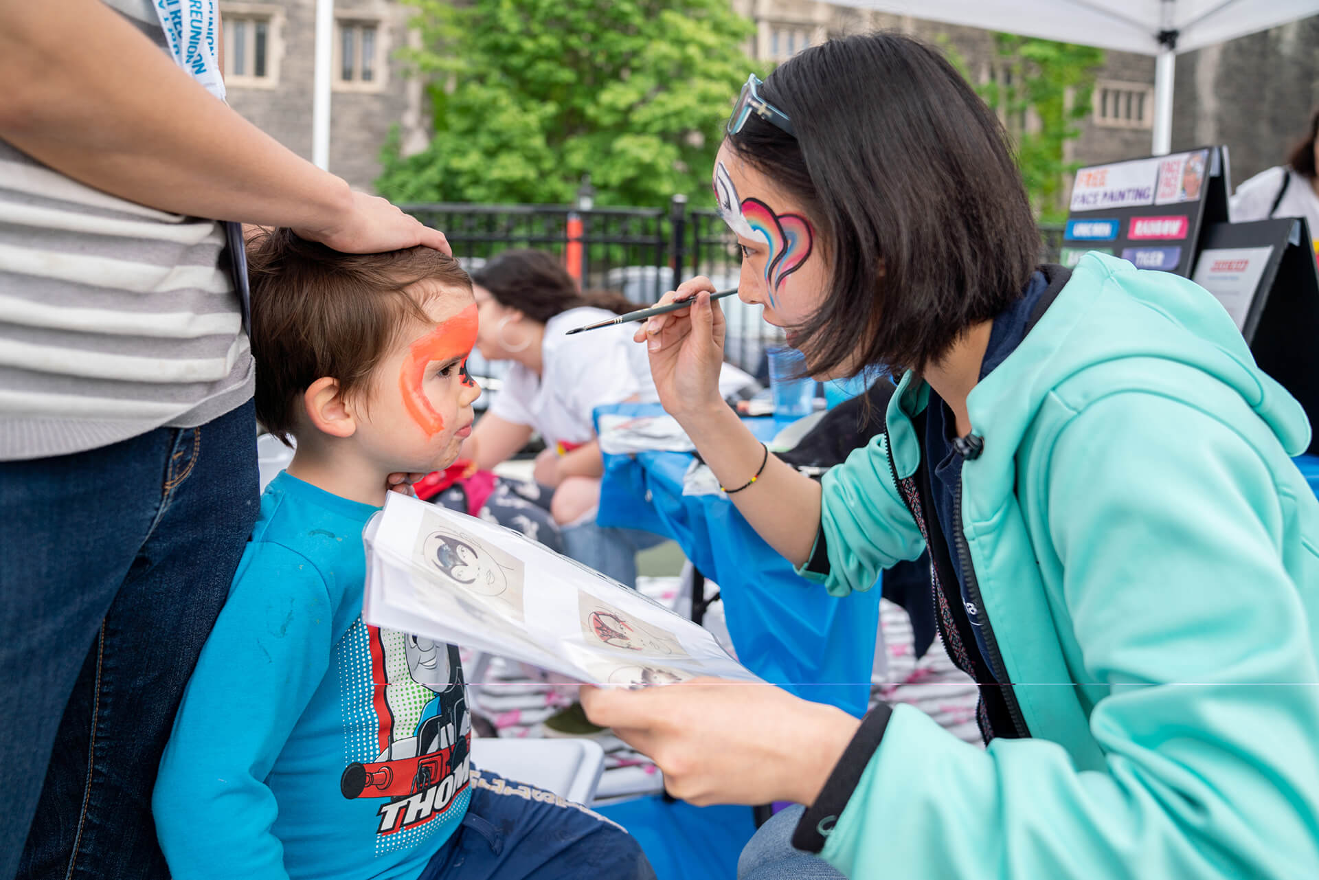 a volunteer with painted face is sitting and painting a kid's face