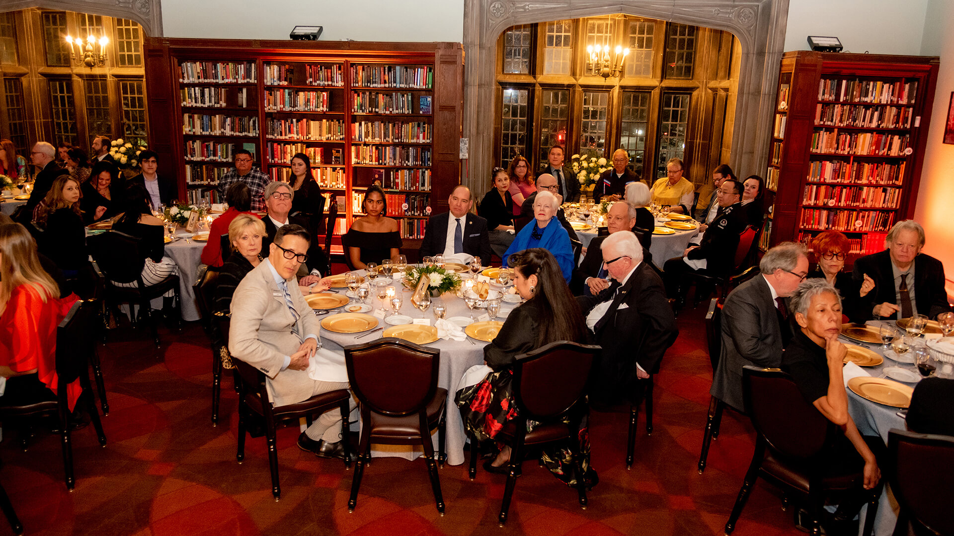 guests are sitting around round dinner tables in a beautiful library surrounding by bookshelves 
