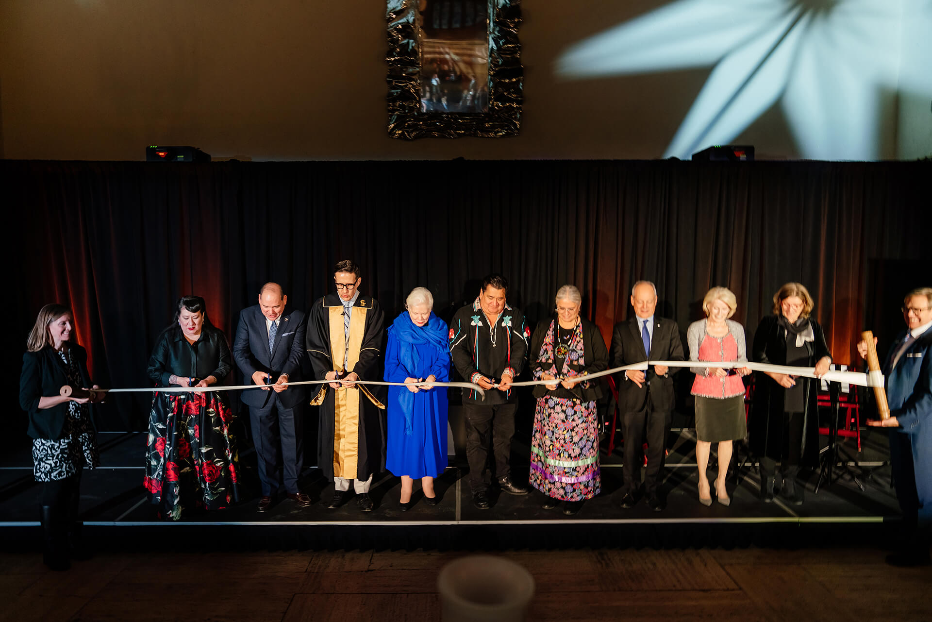 nine people are standing on stage and cutting a long ribbon with scissors which two other people hold on from each side