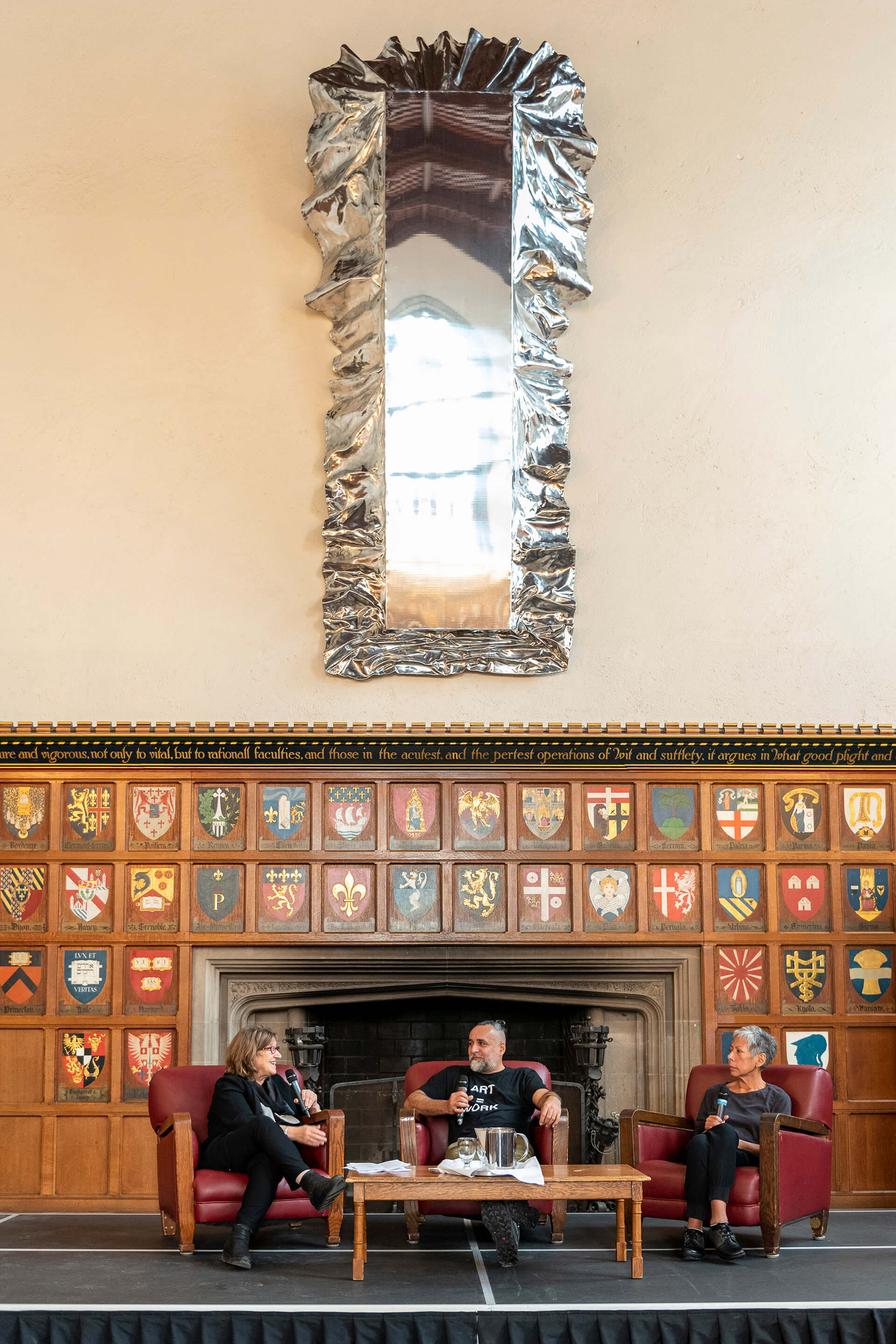 Three people are sitting as a panel and one woman is talking on a mic in front of a fire place and a table is in front of them a large collection of heraldry shields display on the wall behind them and a very tall mirror as artwork on the wall behind