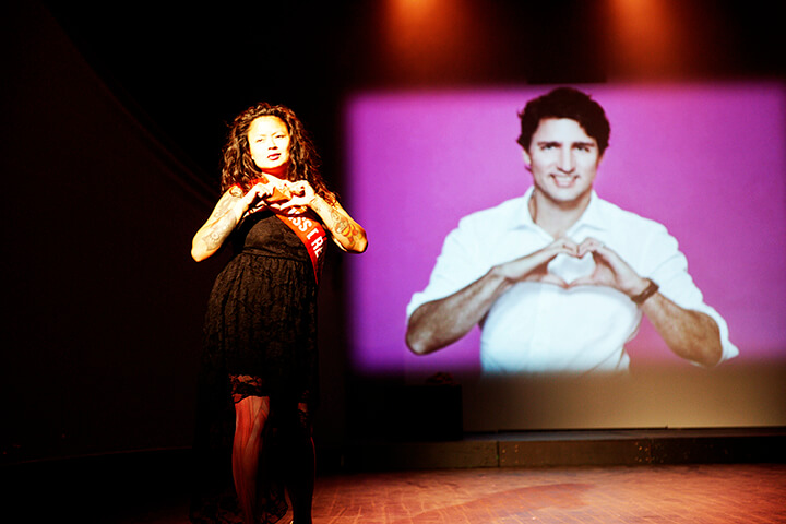 A woman performs on a stage and showing a heart shape with her hands and a screen behind her displays Justin Trudo showing a heart shape with his hands