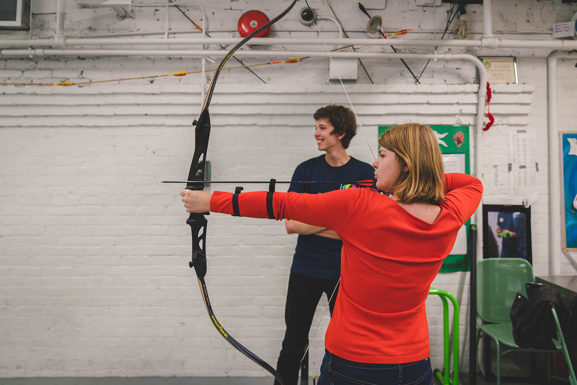 a woman with a bow and arrow trying archery and trying to hit a target and a man volunteer standing beside him in a room