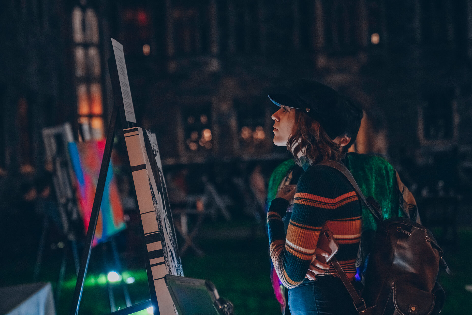 two woman are standing looking at a painting on a tripod on a large quad at night one of them wearing a hat with a backpack and have a glass and a cellphone in her hands
