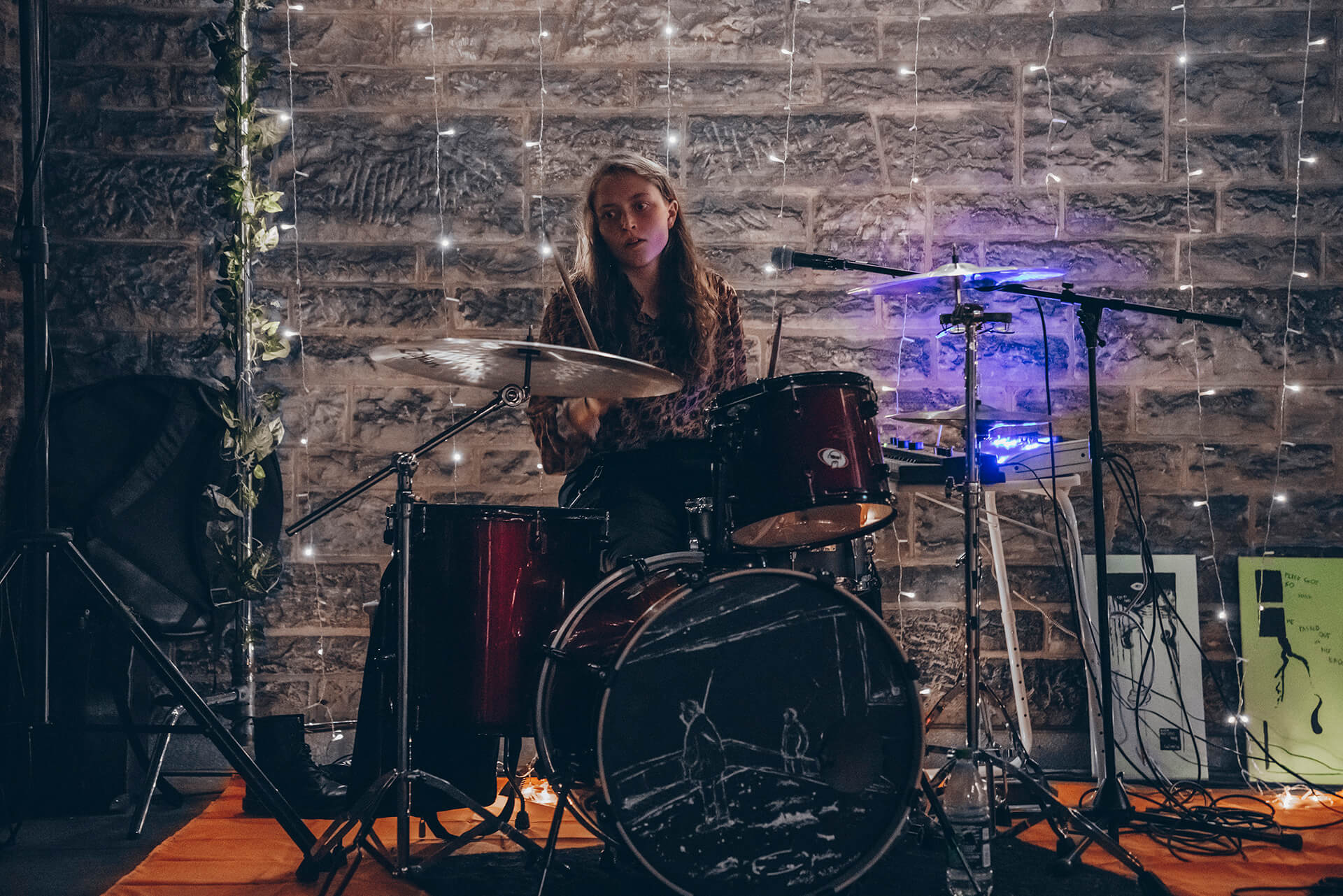 A woman drumming behind a drum at night and some string lights hanging on the wall behind her
