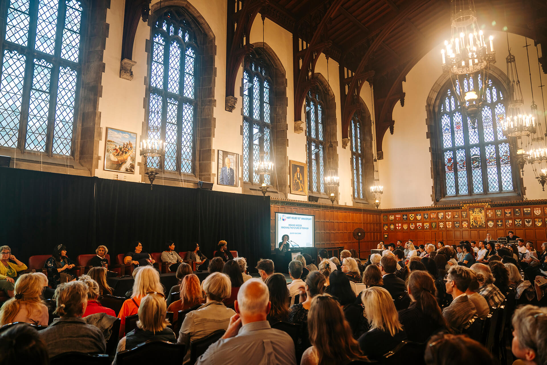 a large group of audience sitting in a great hall with tall arch windows listening to a woman speaker she is behind a podium in front of them a large screen behind the speaker and a panel discussion of seven women sitting in front of audience