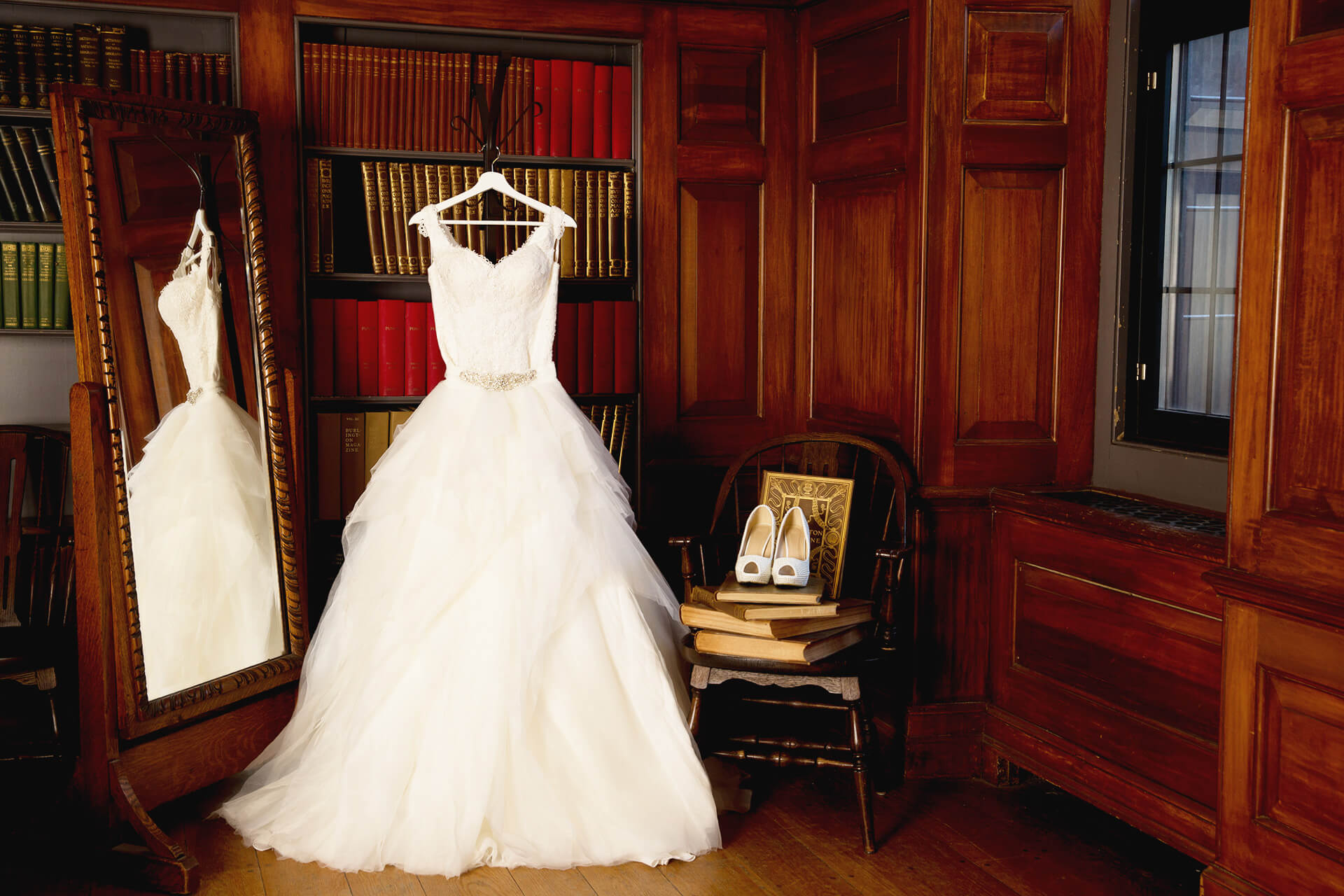 A bridal gown awaits the bride in a room with oak panelling, bookcases, a fireplace, and period tables and chairs.