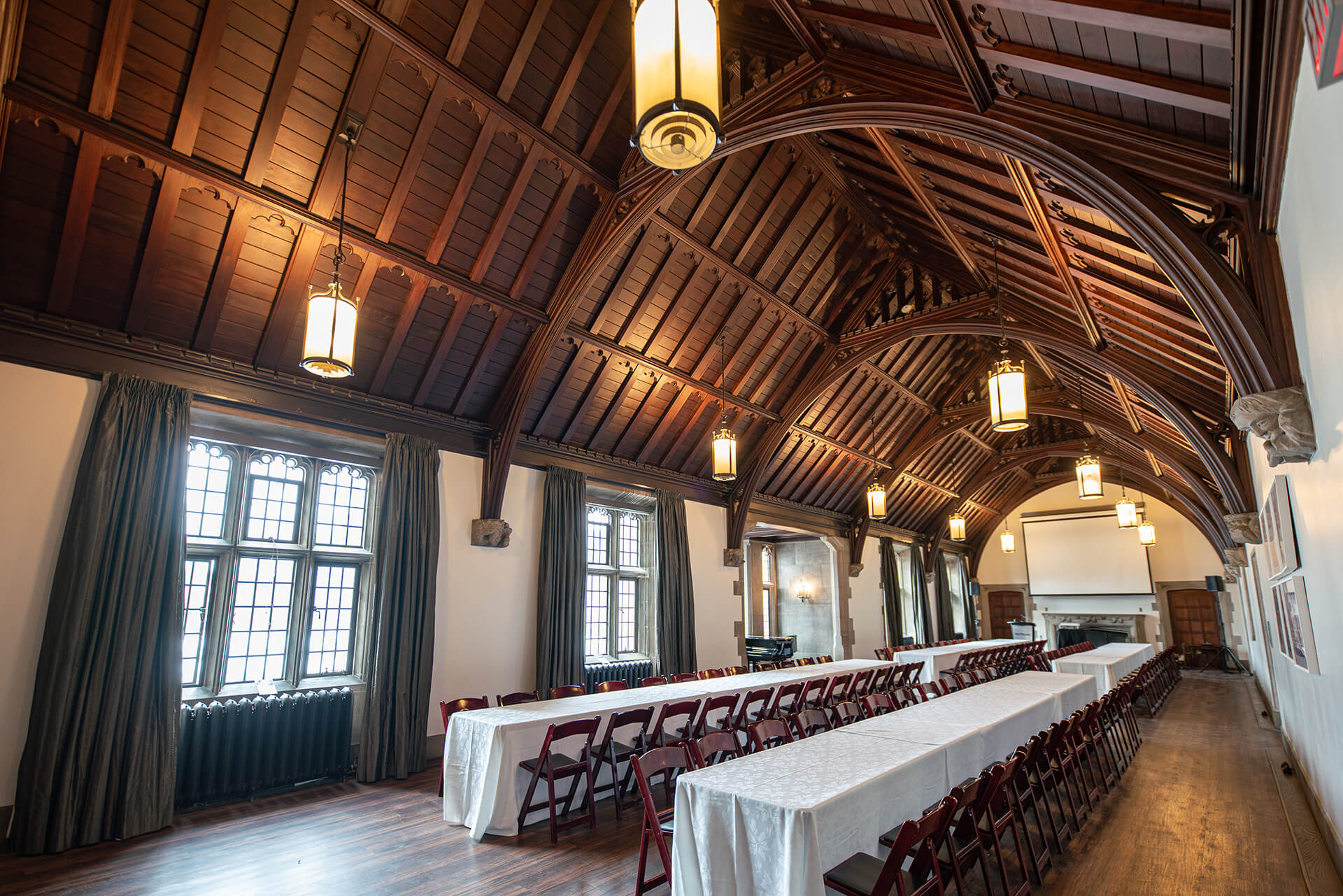 A large room with hardwood floors, gothic windows, vaulted wooden ceiling and tables with seating arranged for an event